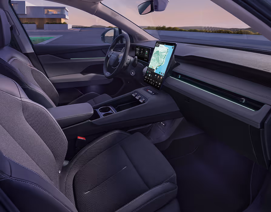 Seitlicher Blick von der Beifahrerseite in das Cockpit des Škoda Peaq mit der dunklen Innenausstattung "Design Selection Loft". Das Interieur zeichnet sich durch graue, strukturierte Sitze, eine breite Mittelkonsole und das markante, hochformatige Infotainment-Display mit aktiver Navigation aus. Entlang der Armaturentafel setzt eine grüne Ambientebeleuchtung im abendlichen Licht frische farbliche Akzente.