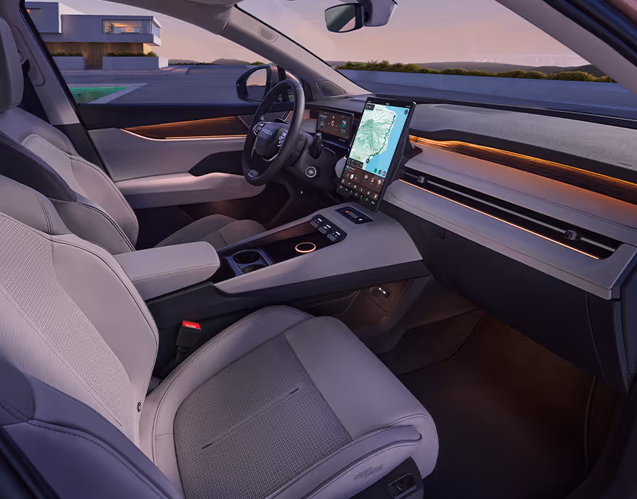 Seitlicher Blick von der Beifahrerseite in das Cockpit des Škoda Peaq mit der hellen Innenausstattung "Design Selection Suite Keramik". Das aufgeräumte Interieur wird von keramikfarbenen Vordersitzen, der breiten Mittelkonsole und dem großen, zentralen Infotainment-Display geprägt. Eine warme, orangefarbene Ambientebeleuchtung durchzieht die Armaturentafel und sorgt im sanften Abendlicht für eine stimmungsvolle Atmosphäre.