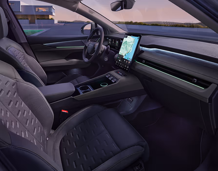 Seitlicher Blick von der Beifahrerseite in das Cockpit des Škoda Peaq mit der zweifarbigen Innenausstattung "Design Selection Lodge". Im Fokus stehen die Vordersitze mit ihren auffällig strukturierten grauen Sitzmittelbahnen, die breite Mittelkonsole und das große, hochformatige Infotainment-Display. Entlang der Armaturentafel und in den Türverkleidungen unterstreicht eine grüne Ambientebeleuchtung die moderne Atmosphäre im sanften Abendlicht.