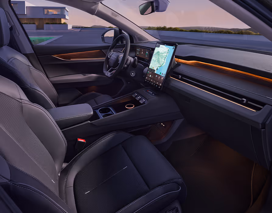Seitlicher Blick von der Beifahrerseite in das Cockpit des Škoda Peaq mit der dunklen Innenausstattung "Design Selection Suite Schwarz". Im Fokus dieser Ansicht stehen die komplett schwarzen Vordersitze, die breite Mittelkonsole sowie das große Infotainment-Display neben dem Multifunktionslenkrad. Eine warme, orangefarbene Ambientebeleuchtung durchzieht die Armaturentafel und setzt elegante farbliche Akzente im abendlichen Interieur.