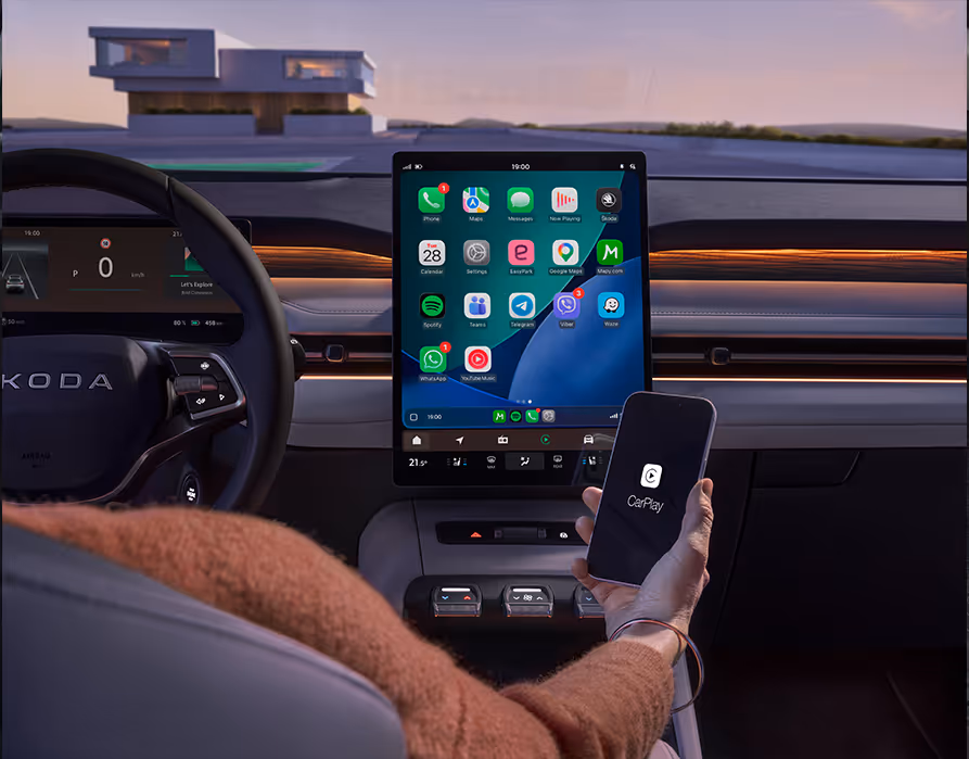 Blick von der Rückbank auf die Armaturentafel des Škoda Peaq. Der Fahrer hält in seiner rechten Hand ein Smartphone mit dem Apple CarPlay-Logo hält. Die App-Oberfläche des Telefons wird dabei übersichtlich auf das große, hochformatige Infotainment-Display in der Mitte gespiegelt. Links daneben sind Teile des Multifunktionslenkrads sowie das digitale Kombiinstrument zu erkennen.