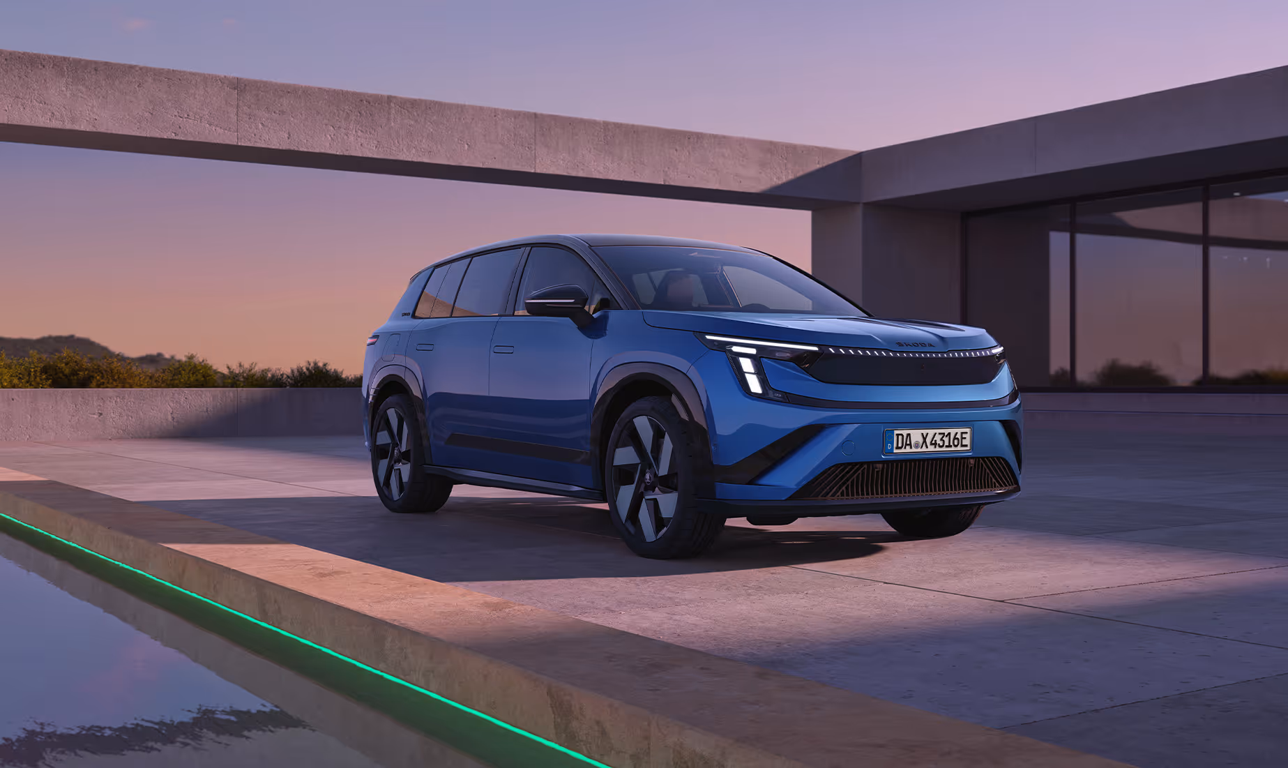 Front- und Seitenansicht des dynamischen Škoda Peaq Sportline in der Lackierung Race-Blau Metallic. Das SUV parkt im sanften Abendlicht vor der klaren, modernen Architektur eines flachen Betongebäudes mit angrenzendem Wasserbecken. Die sportliche Ausrichtung wird durch das markante Frontdesign mit durchgehender Lichtleiste, dunkel abgesetzte Designelemente und große Leichtmetallfelgen unterstrichen.