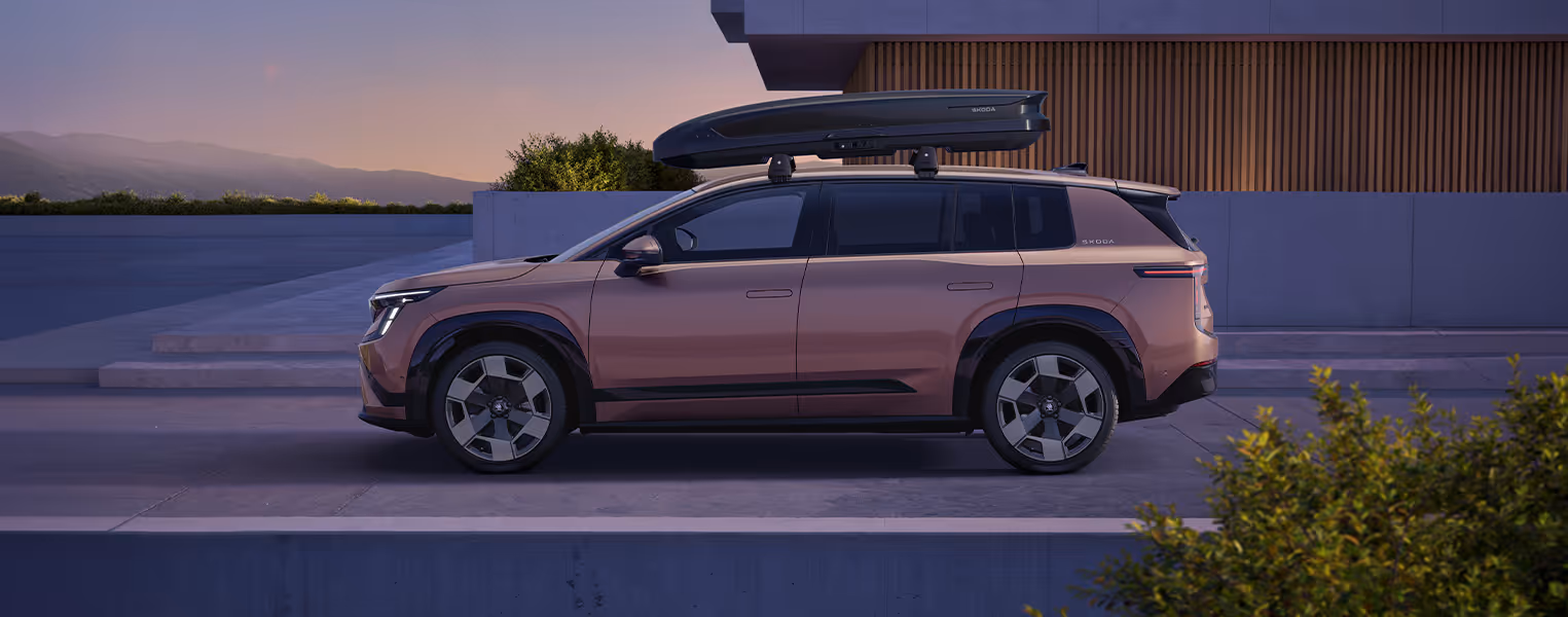 Seitenansicht eines parkenden Škoda Peaq in der Farbe Rosewood-Beige Metallic vor einem modernen Gebäude im sanften Abendlicht. Als praktische Erweiterung des Stauraums ist auf dem Fahrzeug eine große, dunkle Dachbox montiert. 