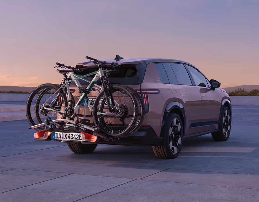 Heck- und Seitenansicht des parkenden Škoda Peaq in der Farbe Rosewood-Beige Metallic im sanften Abendlicht. Am Heck des SUVs ist ein praktischer Fahrradträger montiert, auf dem zwei Fahrräder sicher für den Transport befestigt sind. 