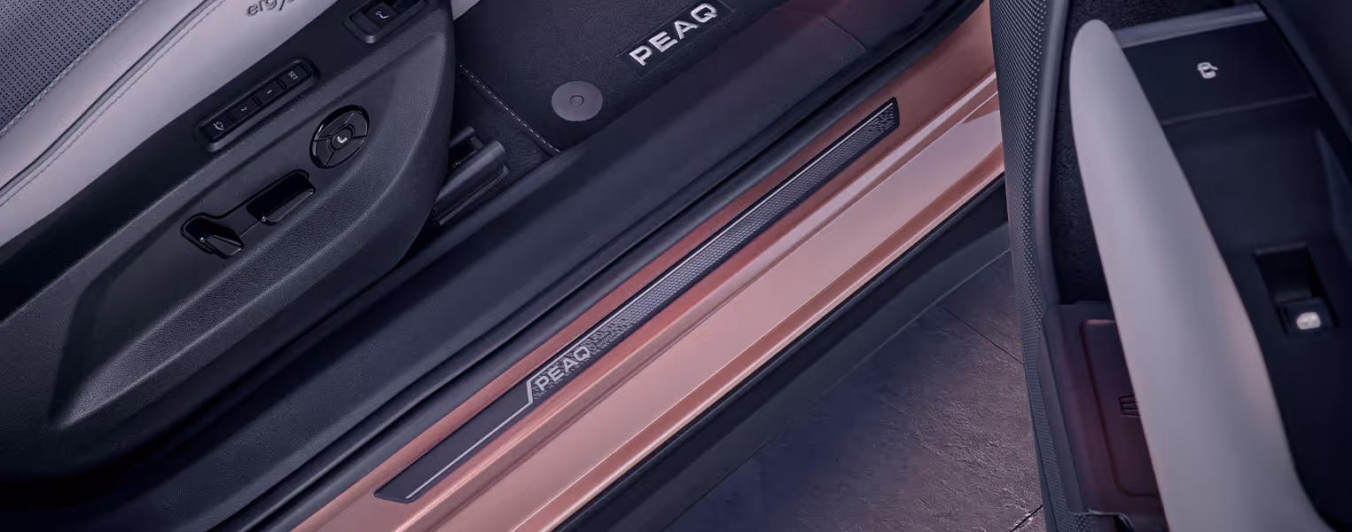 Detailaufnahme des Einstiegsbereichs auf der Beifahrerseite des Škoda Peaq bei geöffneter Tür. Auf dem in Rosewood-Beige Metallic lackierten Türschweller befindet sich eine dunkle Einstiegsleiste, die den markanten Modell-Schriftzug "PEAQ" trägt. 
