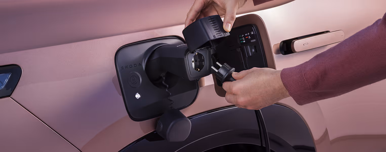 Detailaufnahme des geöffneten Ladeanschlusses am Škoda Peaq in der Lackierung Rosewood-Beige Metallic. Eine Person steckt einen handelsüblichen Stromstecker in einen speziellen Vehicle-to-Load-Adapter, der mit der Ladebuchse des Fahrzeugs verbunden ist.