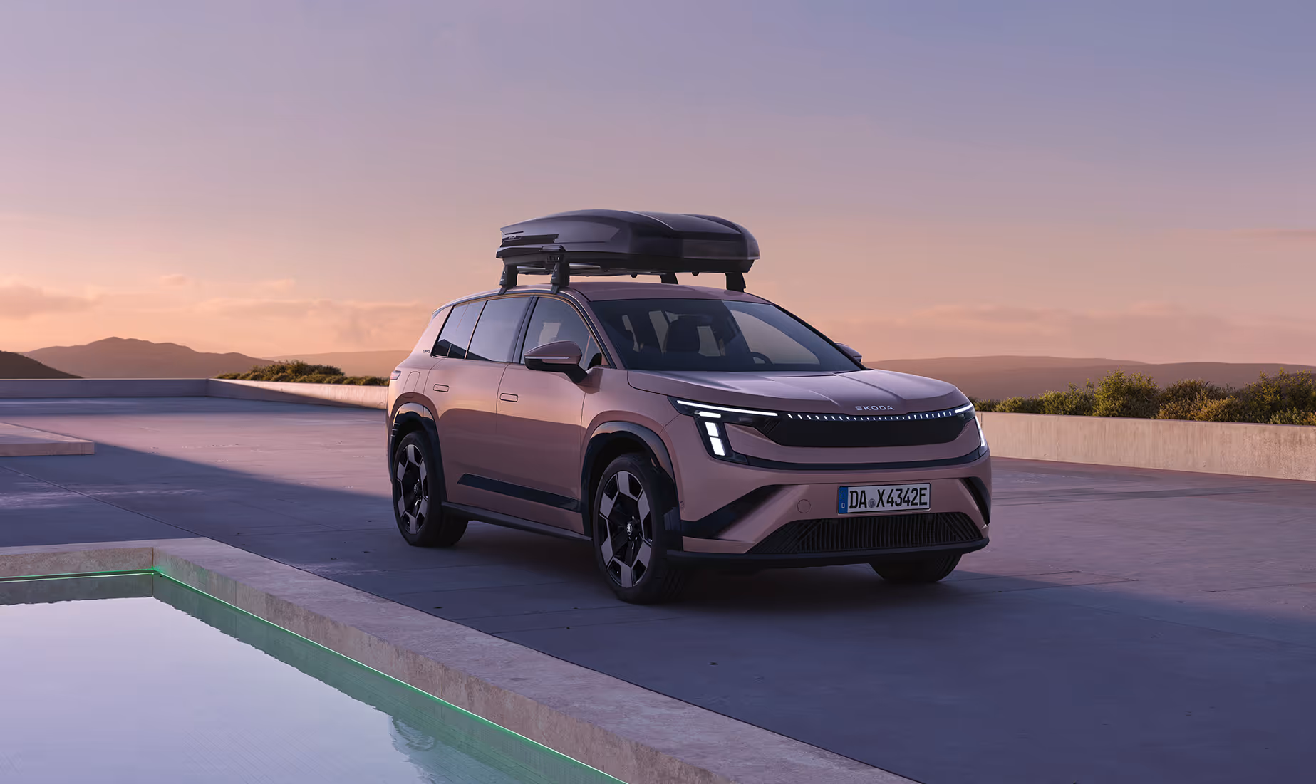 Front- und Seitenansicht des Škoda Peaq in der Lackierung Rosewood-Beige Metallic, der im sanften Abendlicht vor einer modernen Architekturkulisse mit Wasserbecken parkt. Für zusätzlichen Stauraum ist auf dem Fahrzeug eine große, dunkle Dachbox sicher auf Grundträgern montiert.