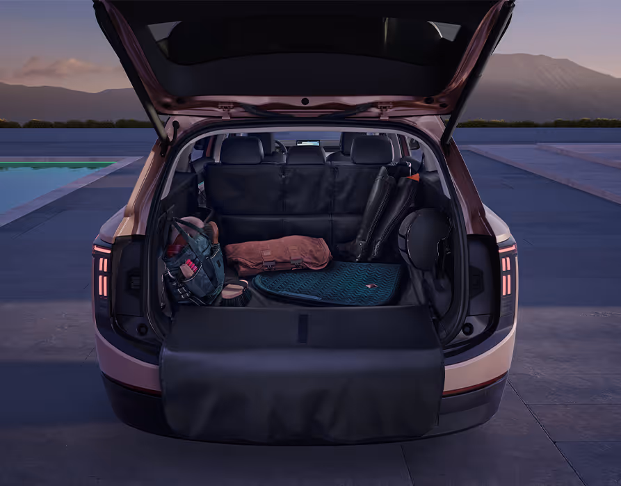 Blick in den geöffneten Kofferraum des Škoda Peaq in der Lackierung Rosewood-Beige Metallic. Der gesamte Gepäckraum ist mit einer dunklen Kofferraumauskleidung geschützt, die auch die Rücksitzlehnen abdeckt und als praktischer Ladekantenschutz über den Heckstoßfänger geklappt ist. Auf der unempfindlichen Unterlage sind verschiedene Ausrüstungsgegenstände wie Taschen, Reitstiefel und ein Helm abgelegt.