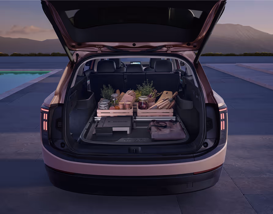 Blick in den geöffneten Kofferraum des Škoda Peaq in der Lackierung Rosewood-Beige Metallic. Der Ladeboden ist mit einer passgenauen, dunklen Kofferraumwanne ausgelegt, die den Innenraum zuverlässig vor Schmutz und Nässe schützt. Darauf stehen rutschfest zwei mit Lebensmitteln gefüllte Holzkisten sowie weiteres Picknick-Zubehör, das durch Cargoelemente sicher an Ort und Stelle gehalten wird.