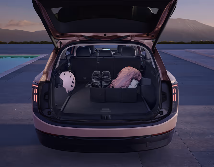 Blick in den geöffneten Kofferraum des Škoda Peaq in der Lackierung Rosewood-Beige Metallic. Auf dem dunklen Ladeboden sorgt eine aufklappbare, schwarze Aufbewahrungsbox für Ordnung, in der ein Paar Stiefel und eine Tasche sicher verstaut sind. Ein seitlich abgelegter Helm unterstreicht das praktische und aufgeräumte Platzangebot für verschiedenste Freizeitaktivitäten.