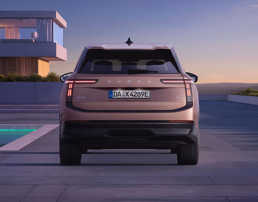 Direkte Heckansicht des Škoda Peaq in der Farbe Rosewood-Beige Metallic, der im sanften Abendlicht vor einer modernen Villa mit Pool parkt. Die Heckpartie zeichnet sich durch schmale, mehrteilige LED-Rückleuchten, einen zentralen Škoda-Schriftzug auf der Kofferraumklappe und einen breit abgesetzten, dunklen Stoßfänger aus.