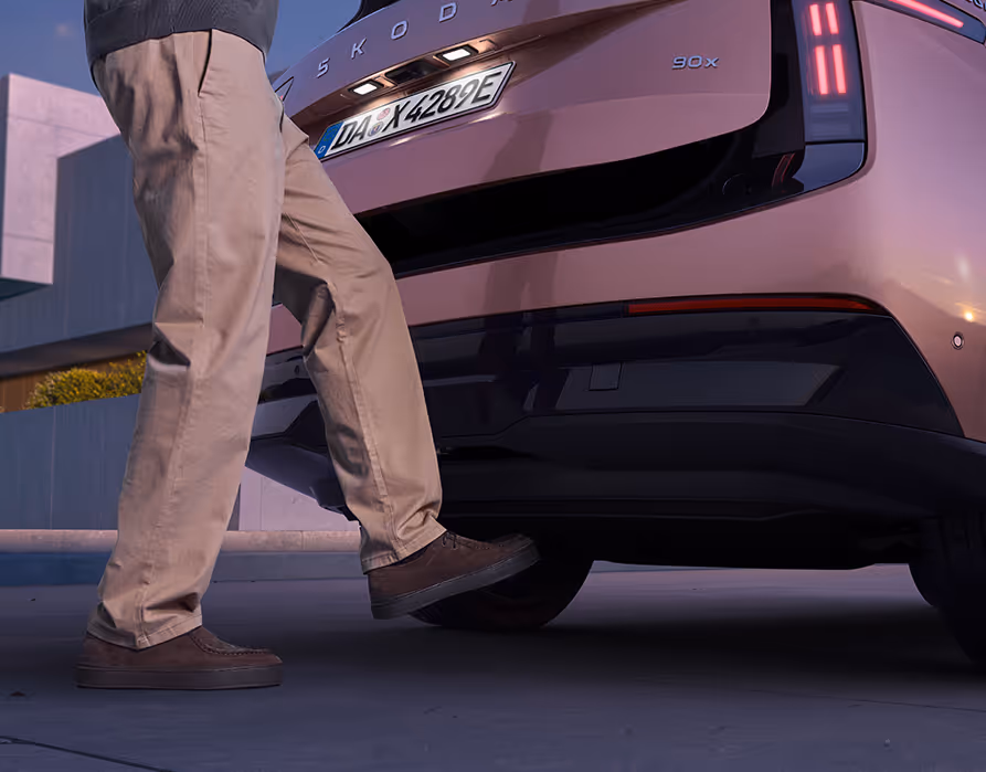 Detailansicht der Heckpartie des Škoda Peaq in der Farbe Rosewood-Beige Metallic. Eine Person demonstriert das berührungslose Öffnen des Kofferraums, indem sie ihren Fuß unter den dunklen Heckstoßfänger bewegt. Daraufhin entriegelt sich die sensorgesteuerte elektrische Heckklappe mit dem beleuchteten Kennzeichen und öffnet sich leicht nach oben.