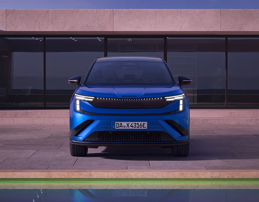 Frontalansicht des Škoda Peaq Sportline in der Lackierung Race-Blau Metallic, der im Abendlicht vor einem modernen Gebäude mit großen dunklen Glasfronten parkt. Das sportliche Frontdesign wird durch eine durchgehende LED-Lichtleiste, eine dunkle Blende und einen markanten Stoßfänger mit tiefen Lufteinlässen hervorgehoben. Im vorderen Bildrand rundet ein flaches Wasserbecken die klare architektonische Szenerie harmonisch ab.
