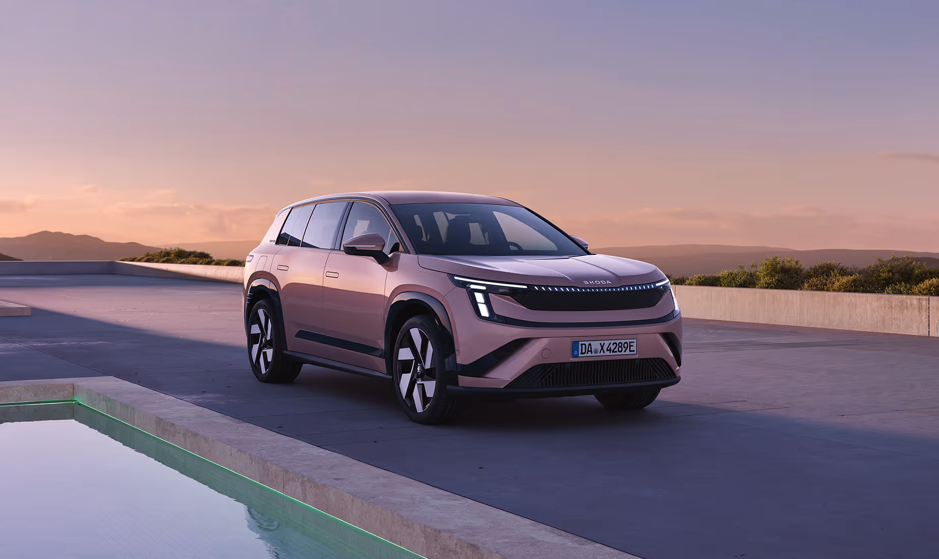 Front- und Seitenansicht des Škoda Peaq in der Lackierung Rosewood-Beige Metallic, der im sanften Abendlicht neben einem flachen Wasserbecken parkt. Die markante Fahrzeugfront wird durch eine durchgehende LED-Lichtleiste, eine dunkle Frontblende und einen breiten unteren Lufteinlass geprägt. Dunkel abgesetzte Radhäuser und aerodynamisch gestaltete Leichtmetallfelgen unterstreichen die klare Silhouette des SUVs vor der ruhigen Landschaftskulisse im Hintergrund.