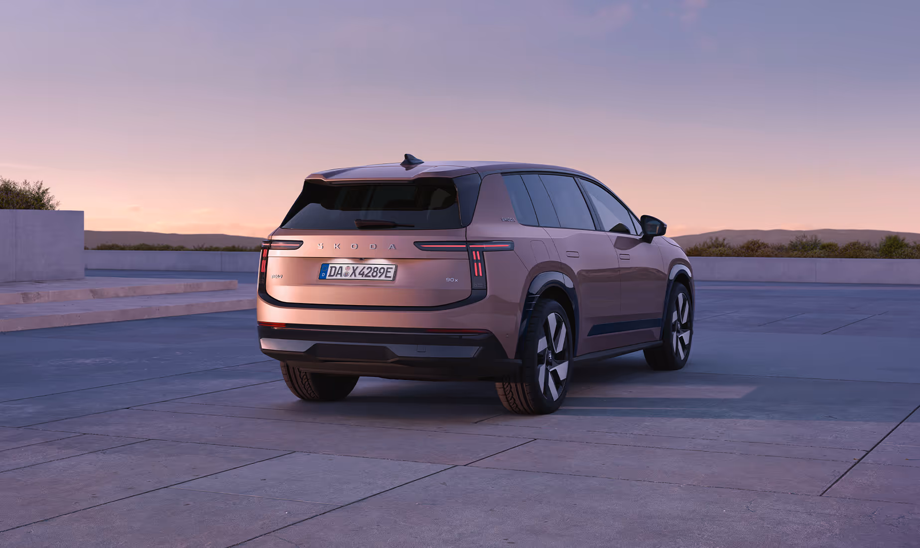 Heck- und rechte Seitenansicht des parkenden Škoda Peaq in der Lackierung Rosewood-Beige Metallic unter einem stimmungsvollen Abendhimmel. Die markante Heckpartie zeichnet sich durch schmale LED-Rückleuchten, einen dezenten Dachspoiler und den zentralen Škoda-Schriftzug auf der Kofferraumklappe aus. Dunkel abgesetzte Radhäuser und aerodynamische Leichtmetallfelgen unterstreichen die klare Linienführung des SUVs vor der ruhigen Landschaftskulisse im Hintergrund.
