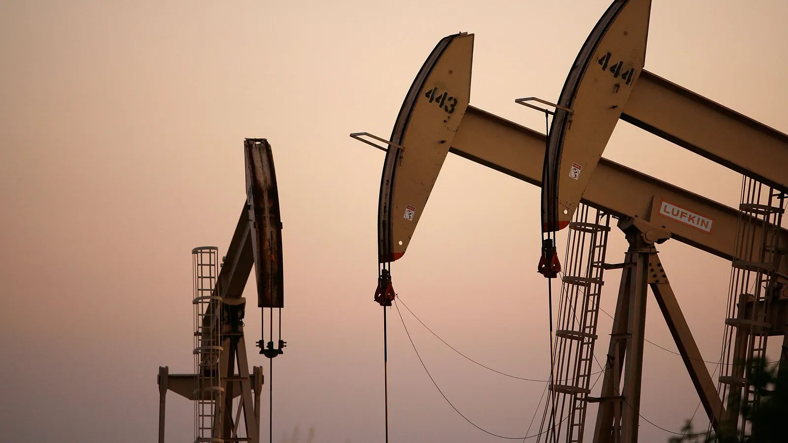 A wide shot of three large oil pumpjacks set against a hazy, warm orange and beige sky during twilight or sunset. The pumpjack in the foreground on the right features the number "444" and the brand name "LUFKIN" on its side, while the middle one is numbered "443." The pumpjacks are positioned at an angle, showing their mechanical arms, ladders, and cables, with a third, weathered pumpjack visible slightly further in the background on the left.