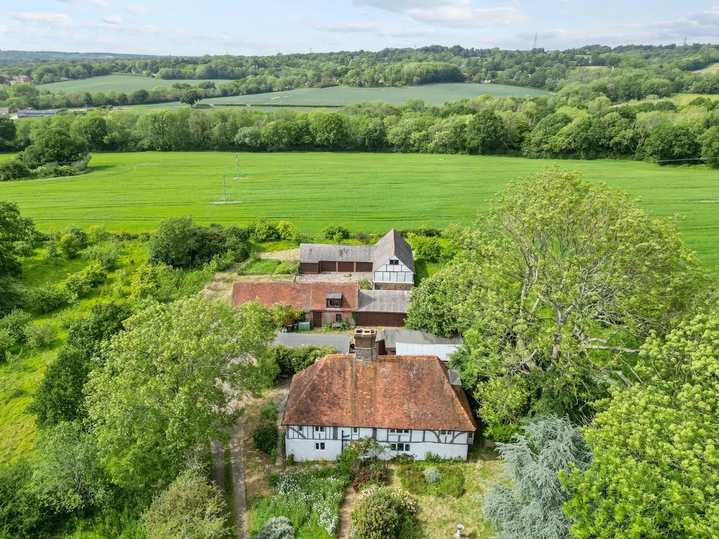 Aerial view of countryside with a farmhouse, outbuildings, trees, and surrounding green fields.