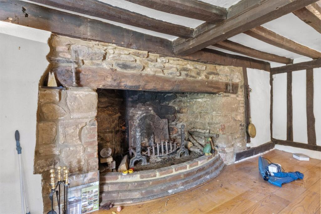 Large rustic stone fireplace with wooden beam mantel and firewood inside, in a room with wooden floor and beams.