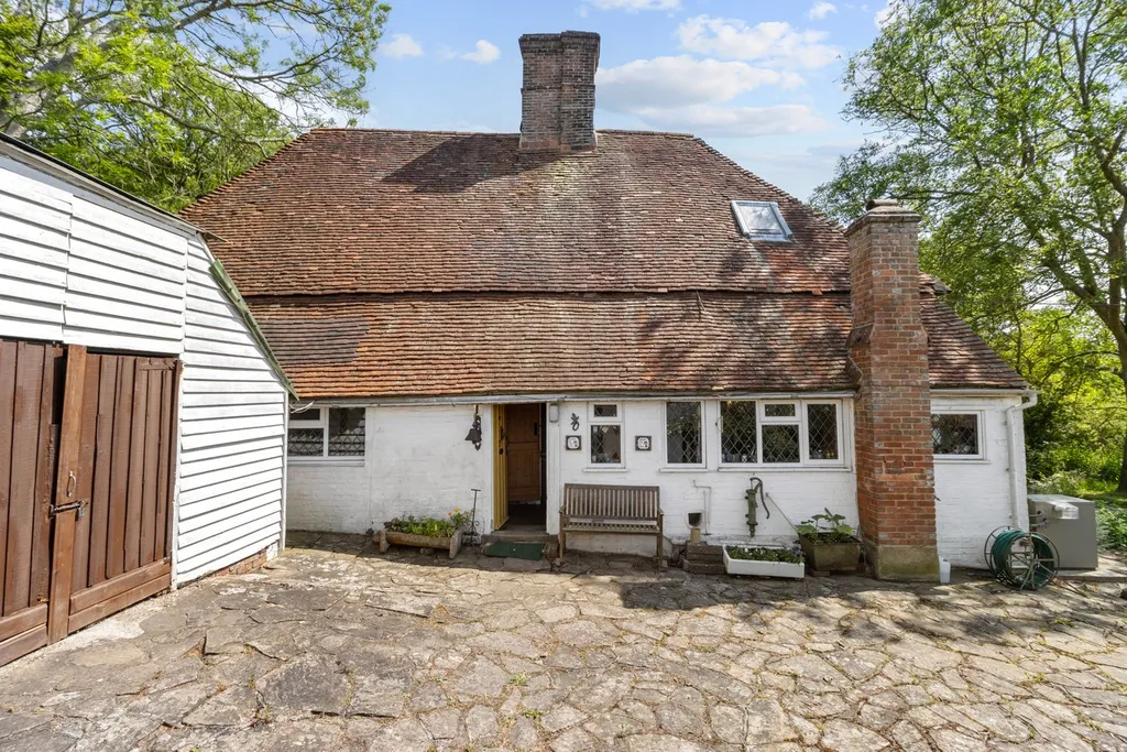 Front of a rustic white cottage with a brown tiled roof, chimney, wooden bench, and stone driveway.