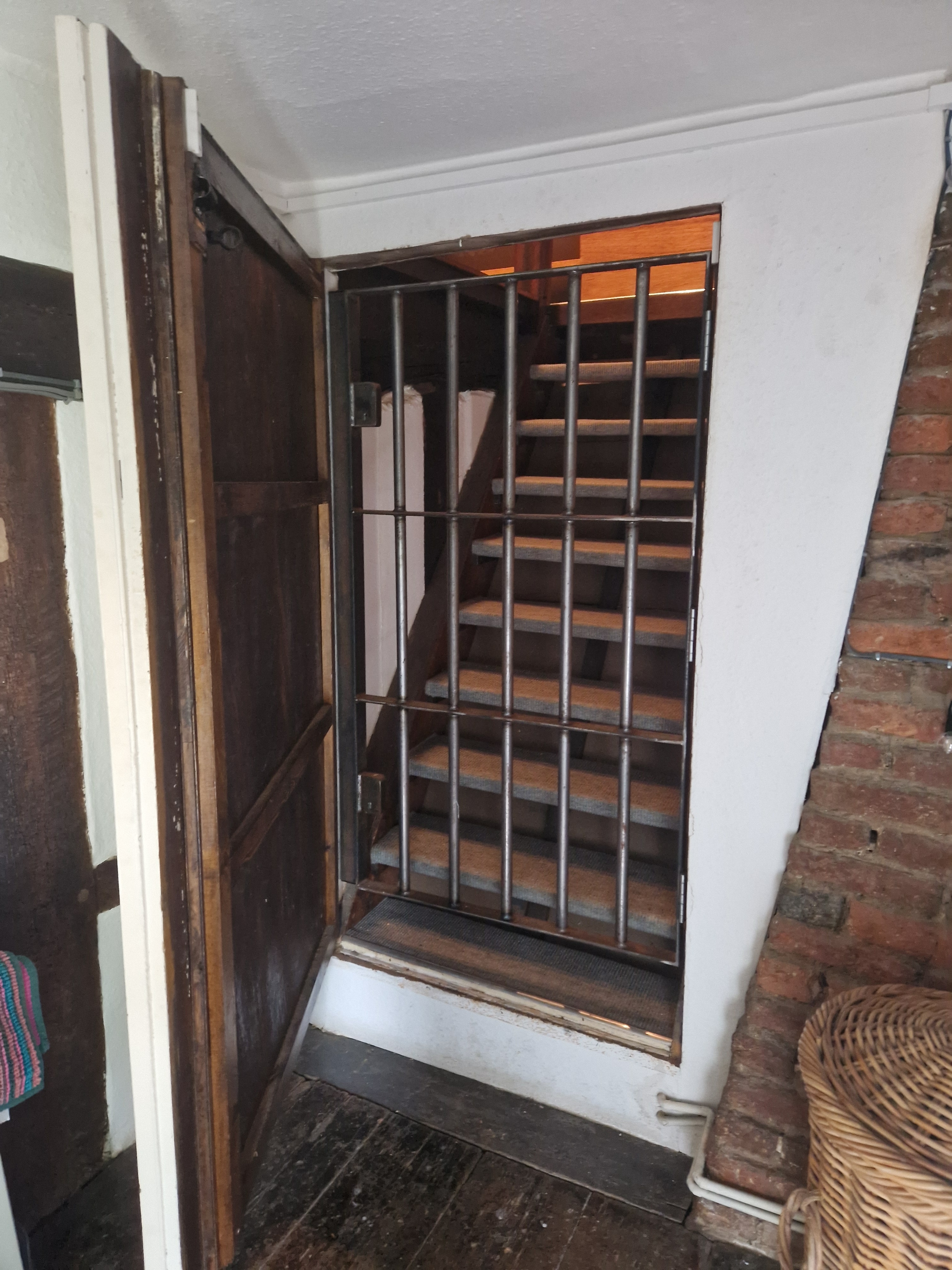 Open wooden door and metal gate reveal staircase inside a rustic interior with brick wall and wooden floor.