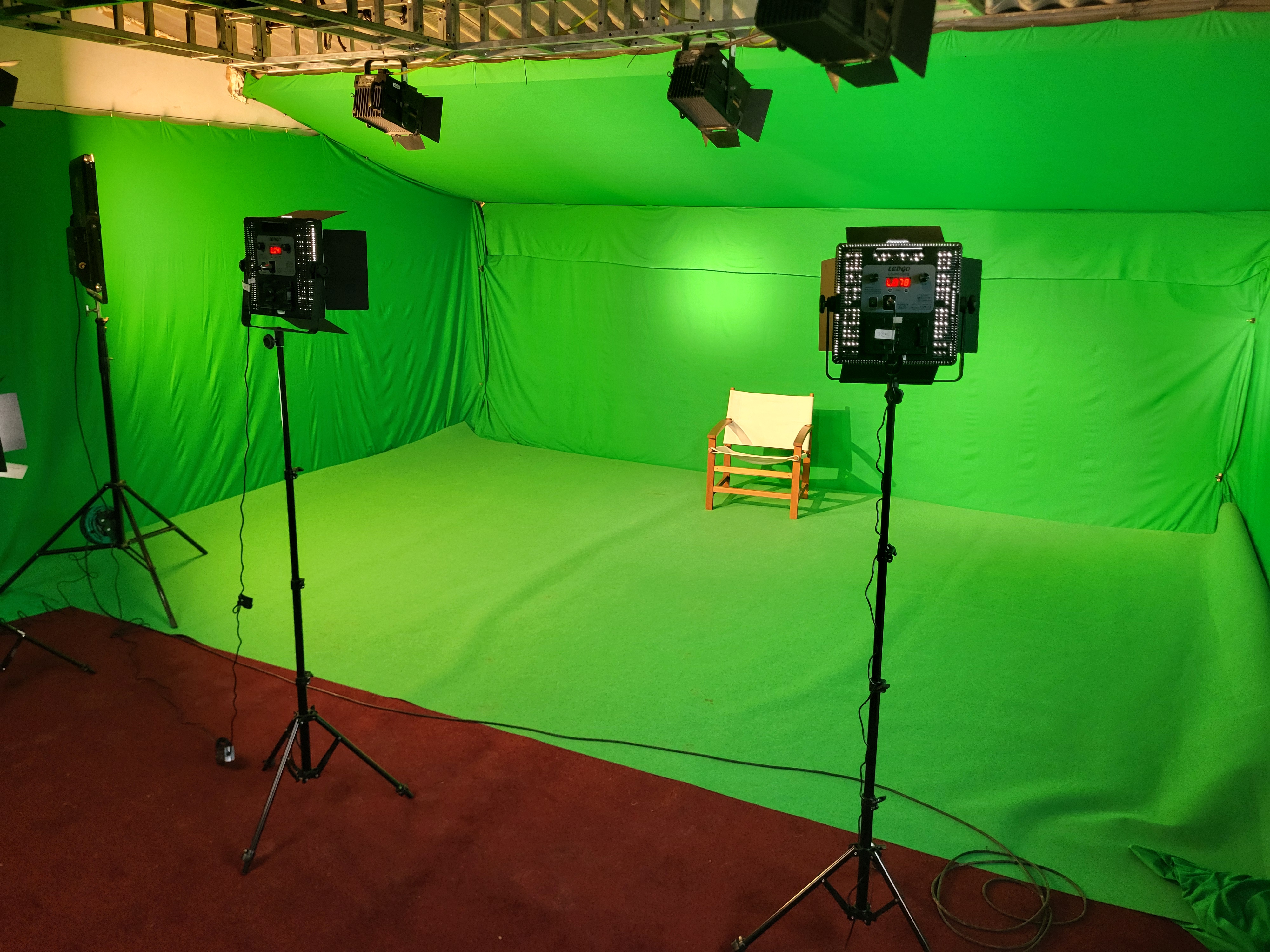 Empty green screen studio with three lighting stands and a wooden chair with cloth seat.