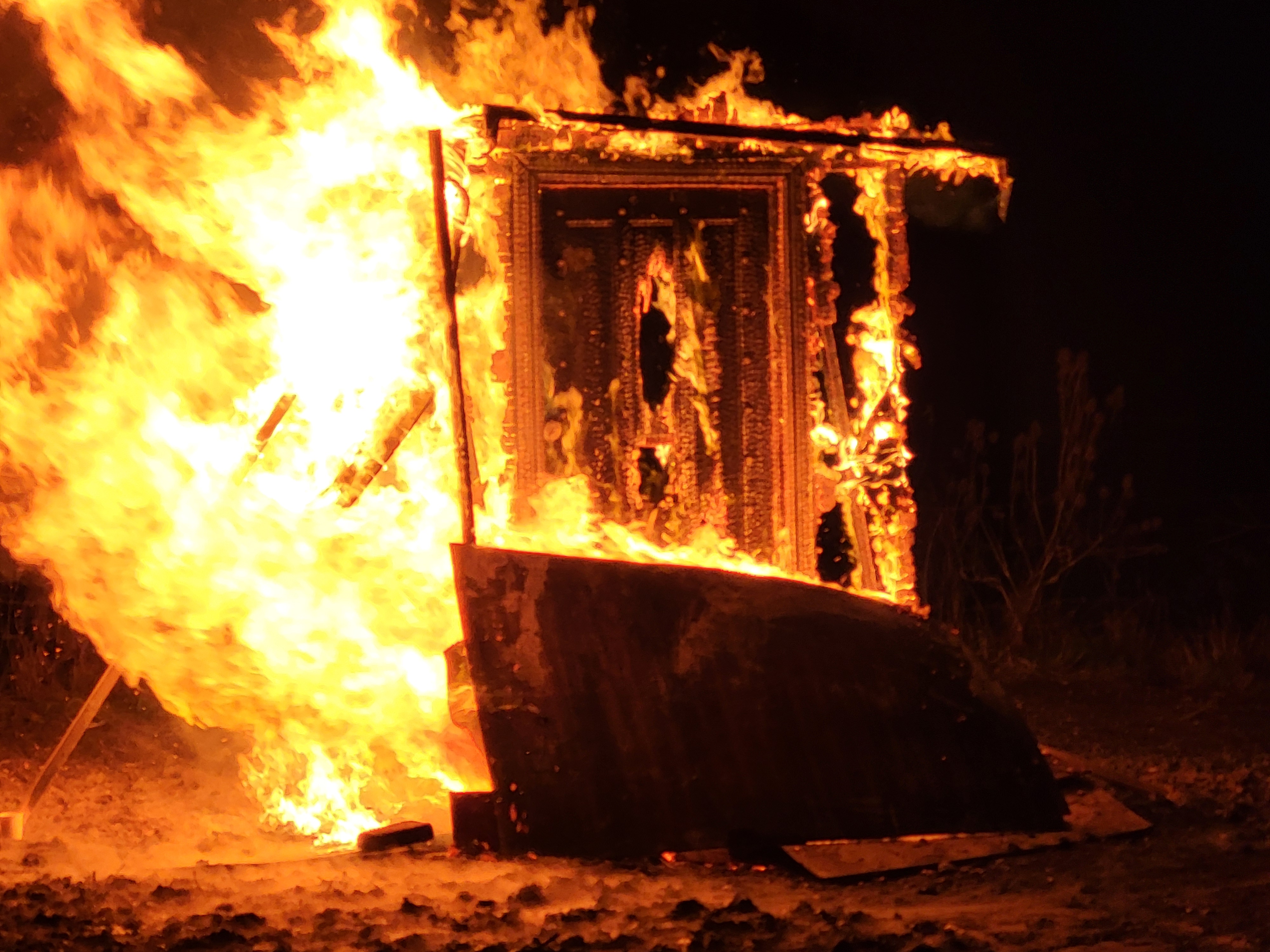 Large wooden structure engulfed in bright orange flames at night on dirt ground.