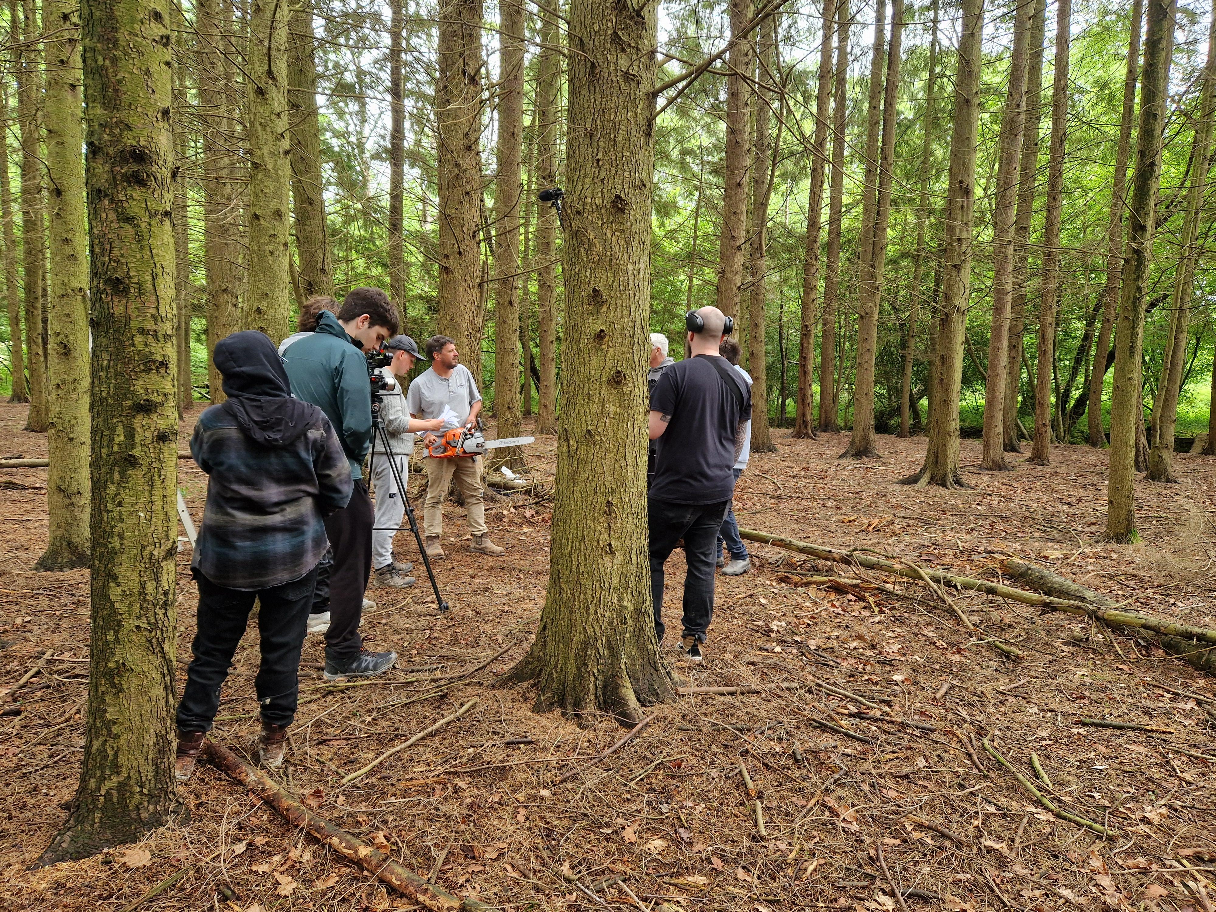 Group of people filming a man holding a chainsaw in a dense forest with tall, thin trees.