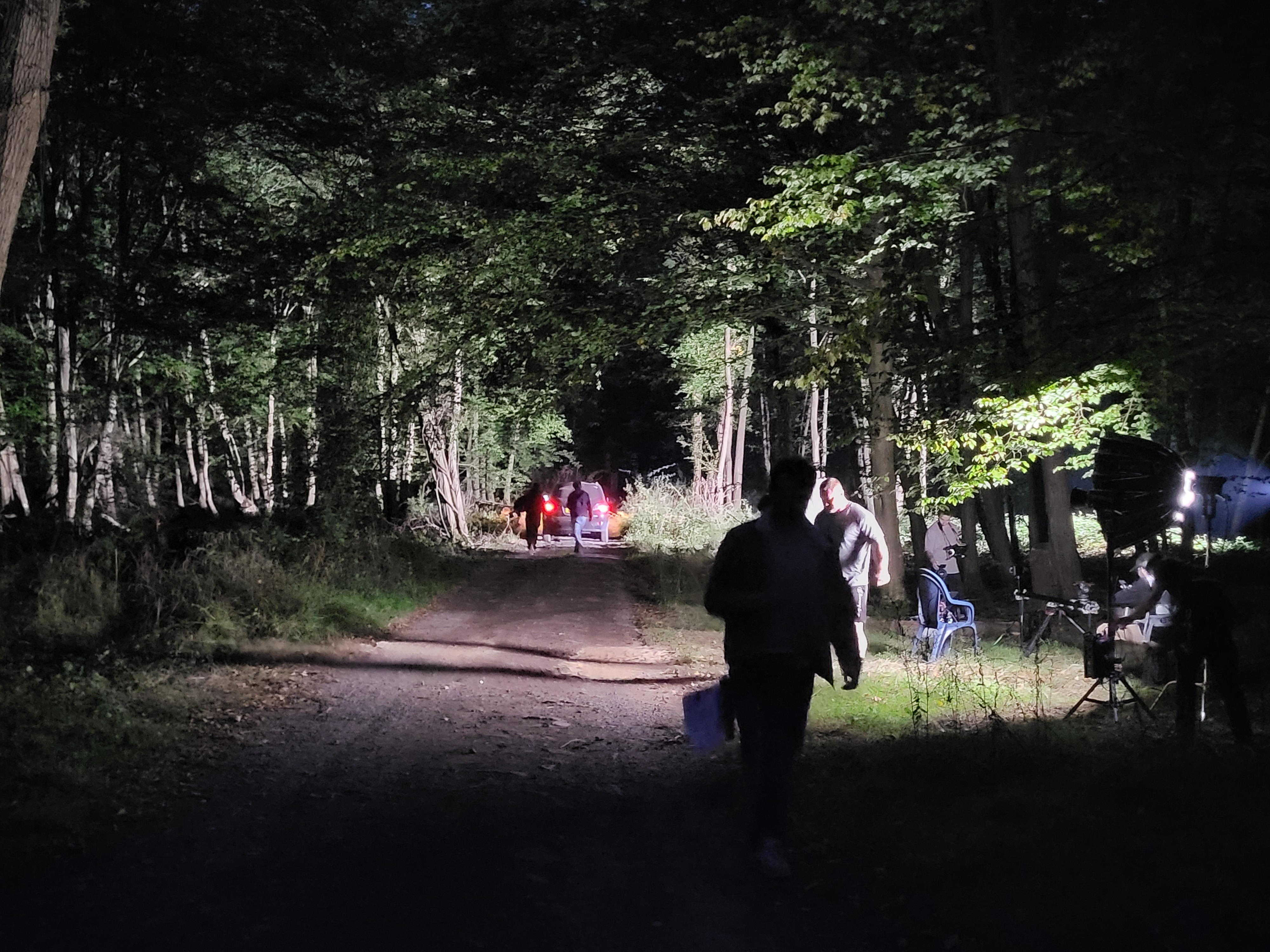 Night forest scene with people, car headlights, and a light setup with crew on the right.