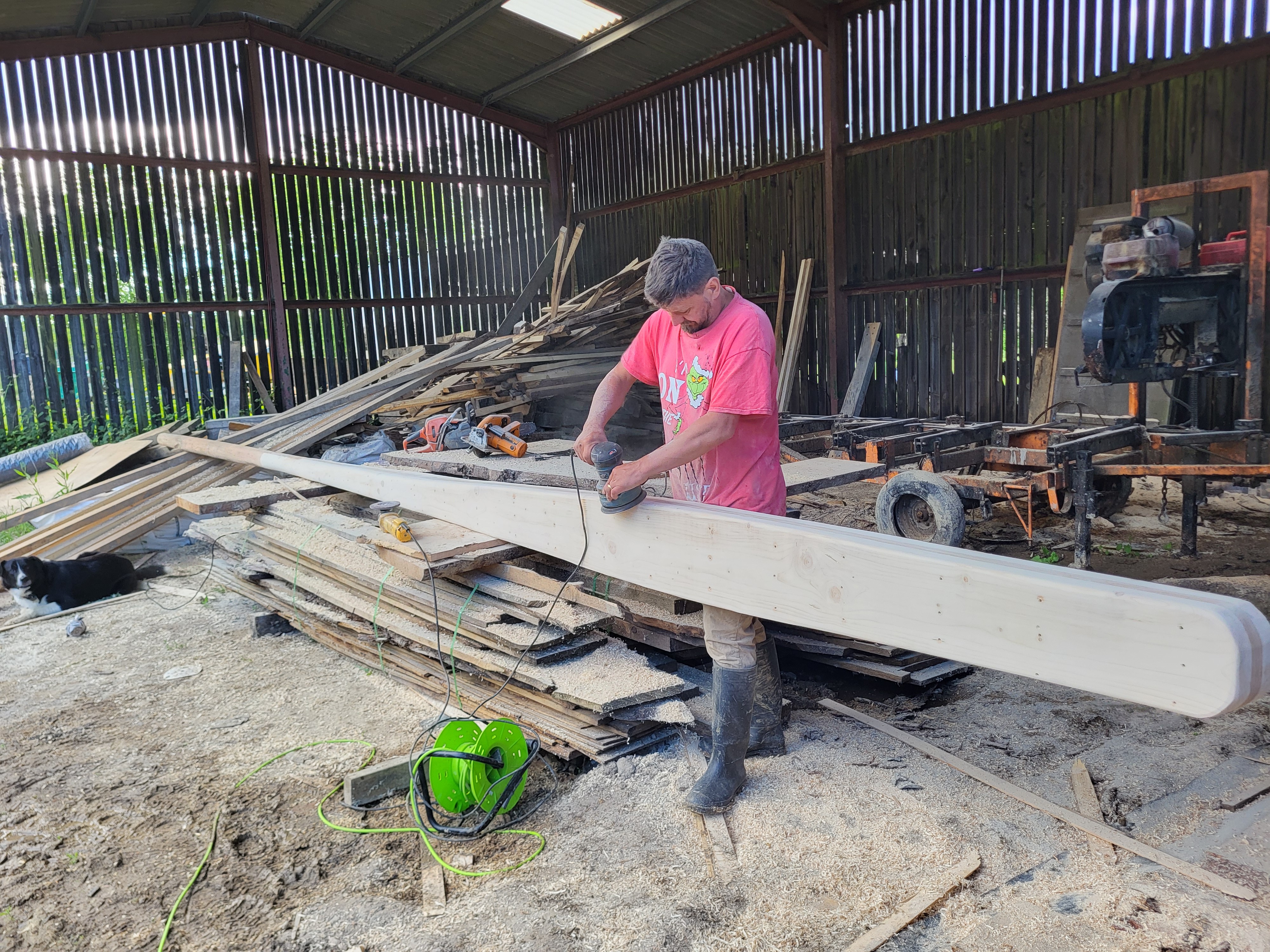Man in pink shirt sanding a long wooden beam in a wood workshop with stacked lumber and tools nearby.