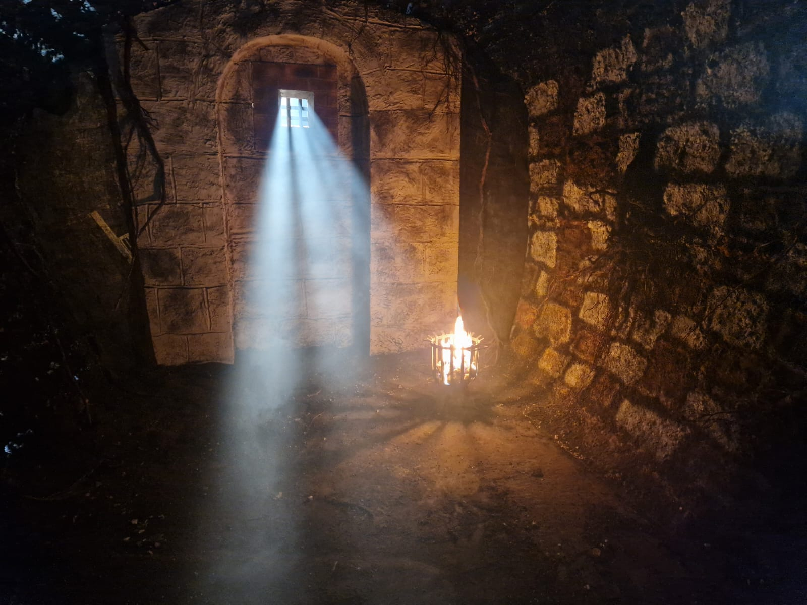 Sunlight beams through a barred window into a stone room with a lit fire basket on the floor.