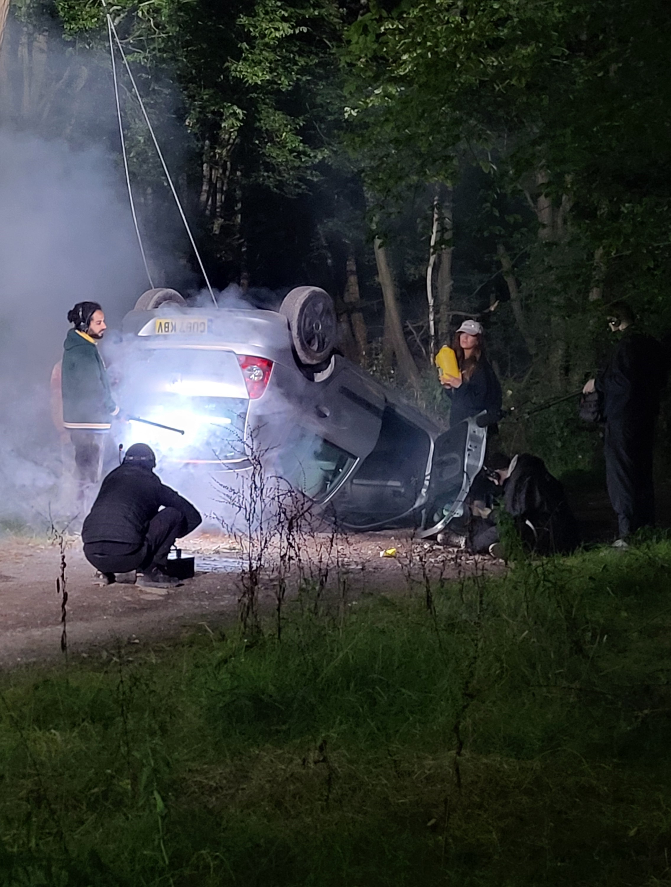 Overturned car on dirt road surrounded by people with smoke and trees in the background at night.