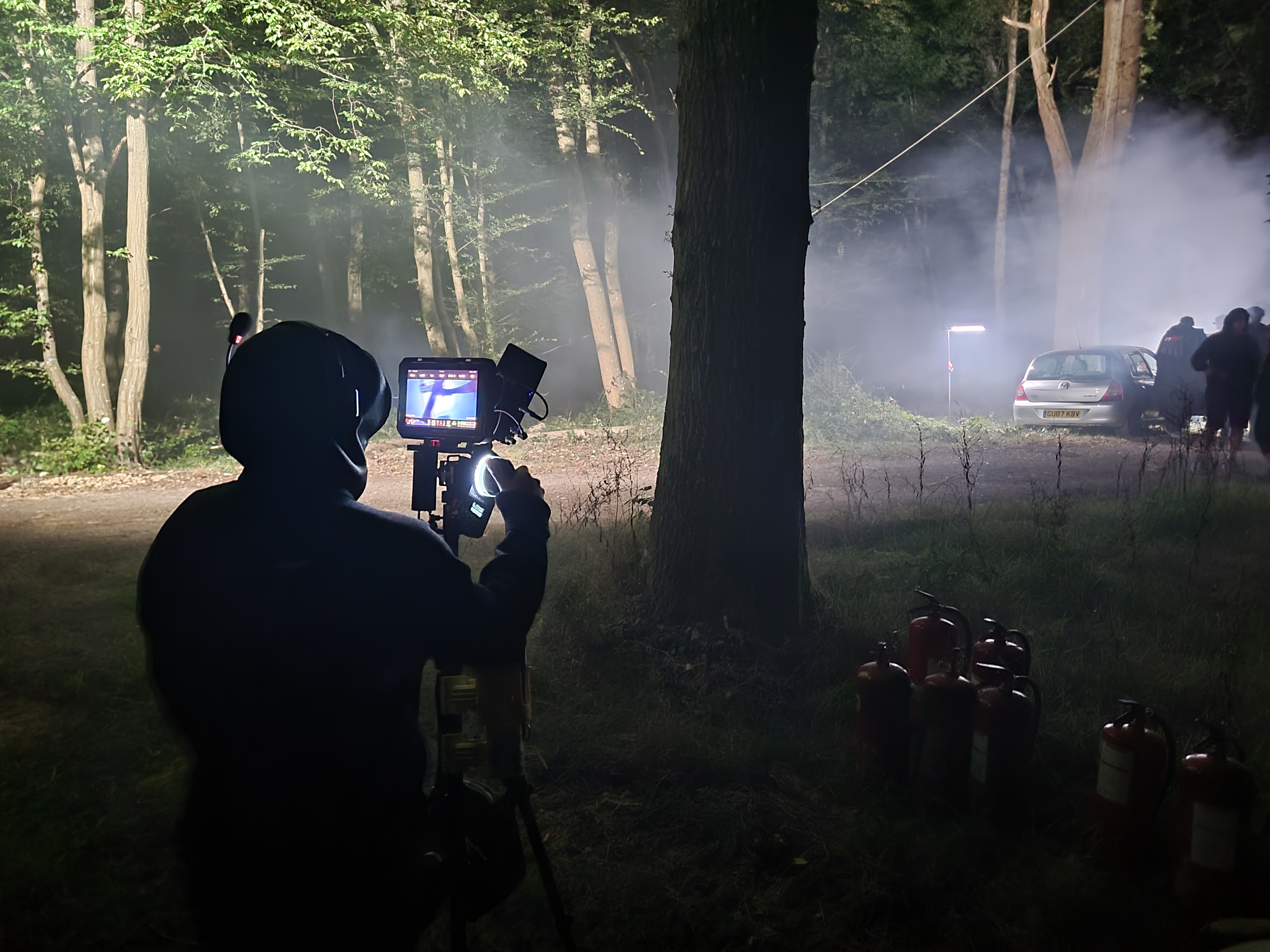 Person filming a foggy forest scene at night with camera on tripod and parked cars in background.