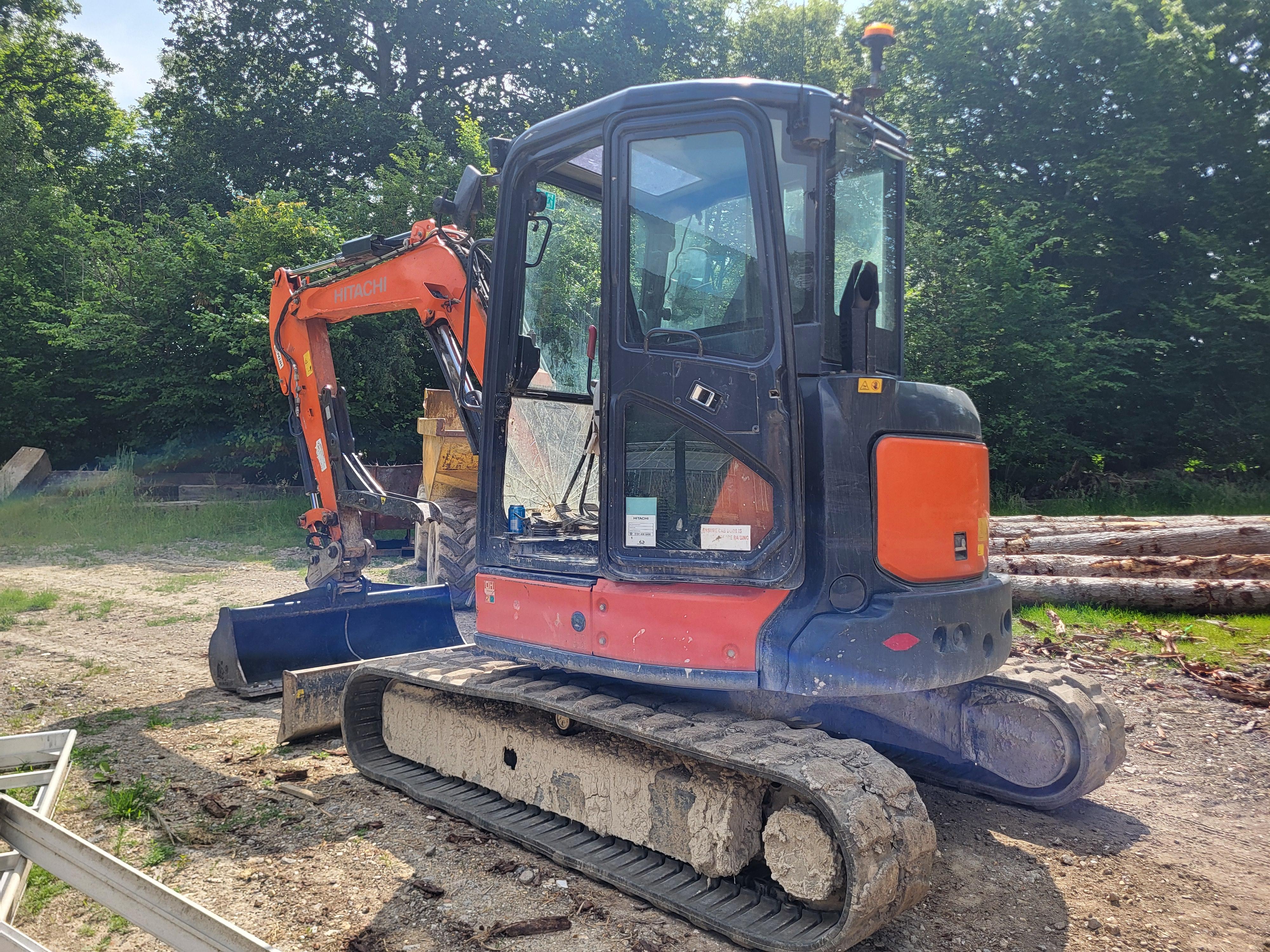 Orange and black Hitachi excavator with dirt-covered tracks parked on gravel near logs and trees.