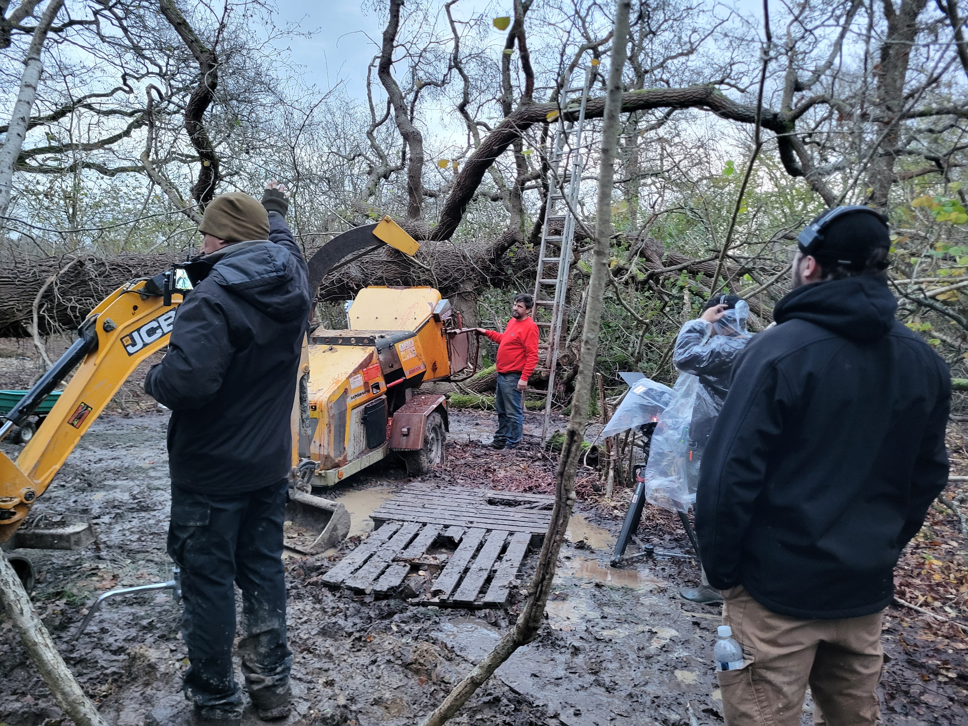Four men in a muddy forest clearing near a yellow wood chipper and ladder by fallen trees.