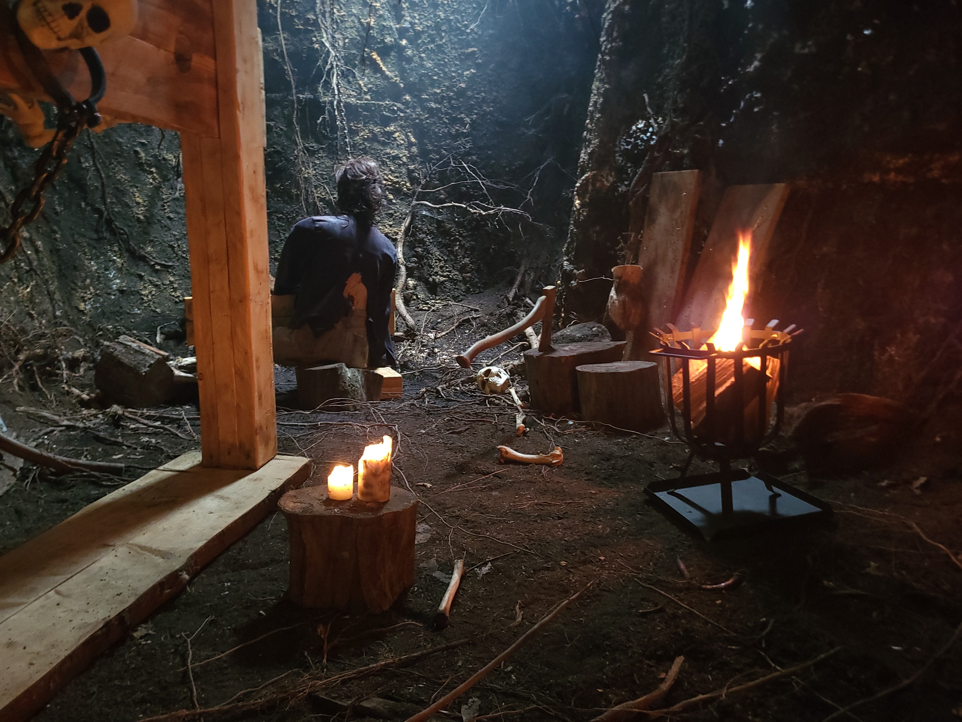 Dark cave setting with a seated figure, candles on a tree stump, skull, bones, and a fire in a metal brazier.