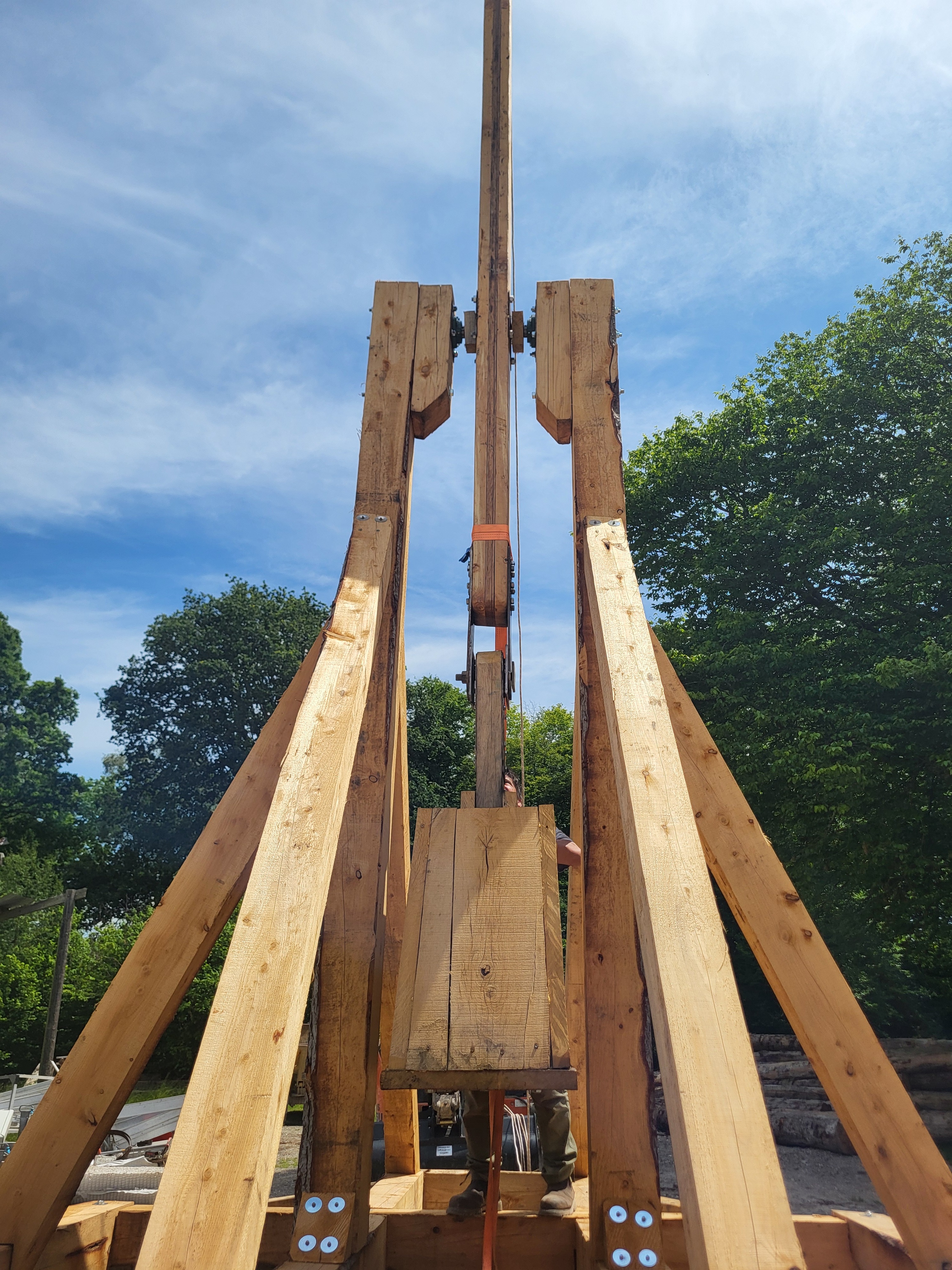 Large wooden trebuchet catapult with a person standing behind it outdoors on a sunny day.