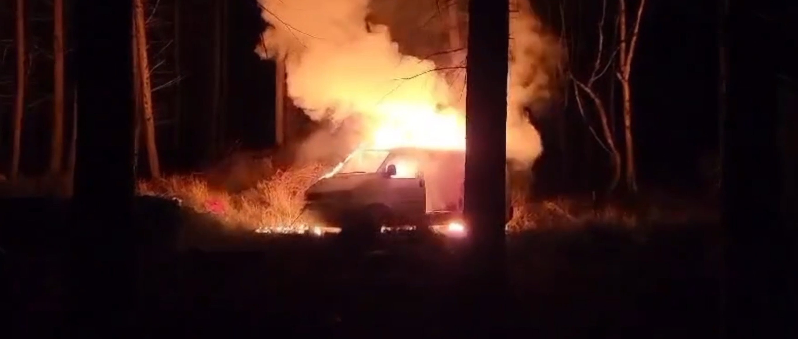 Van engulfed in flames and smoke at night in a wooded area.