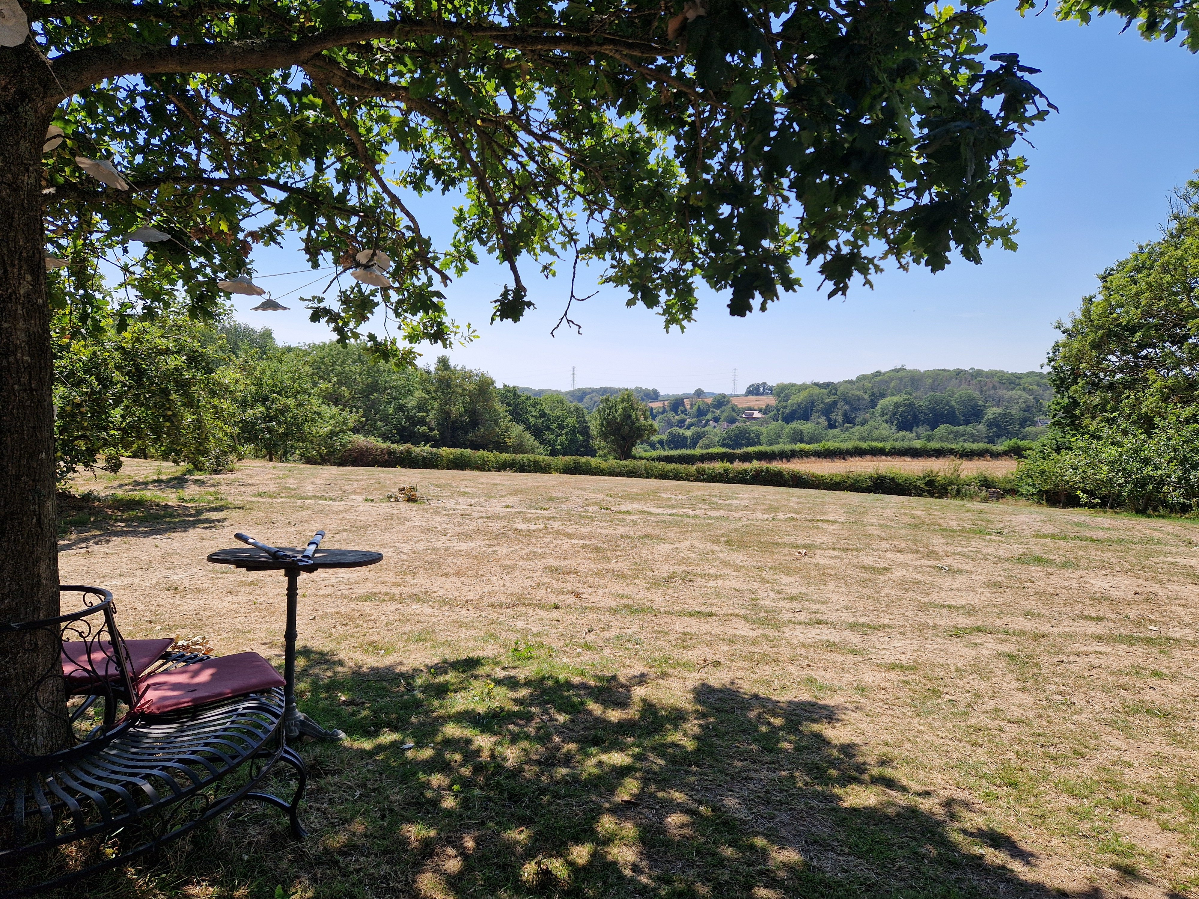 Shaded metal bench and table under a tree overlooking a dry grassy field and distant hills.