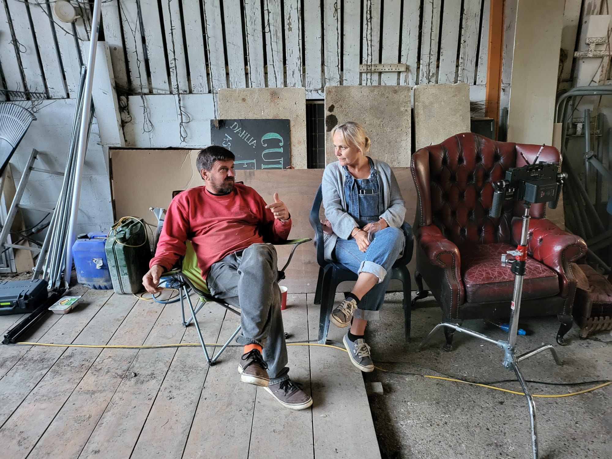 Man and woman seated in a rustic room engaged in conversation with equipment and chairs around them.