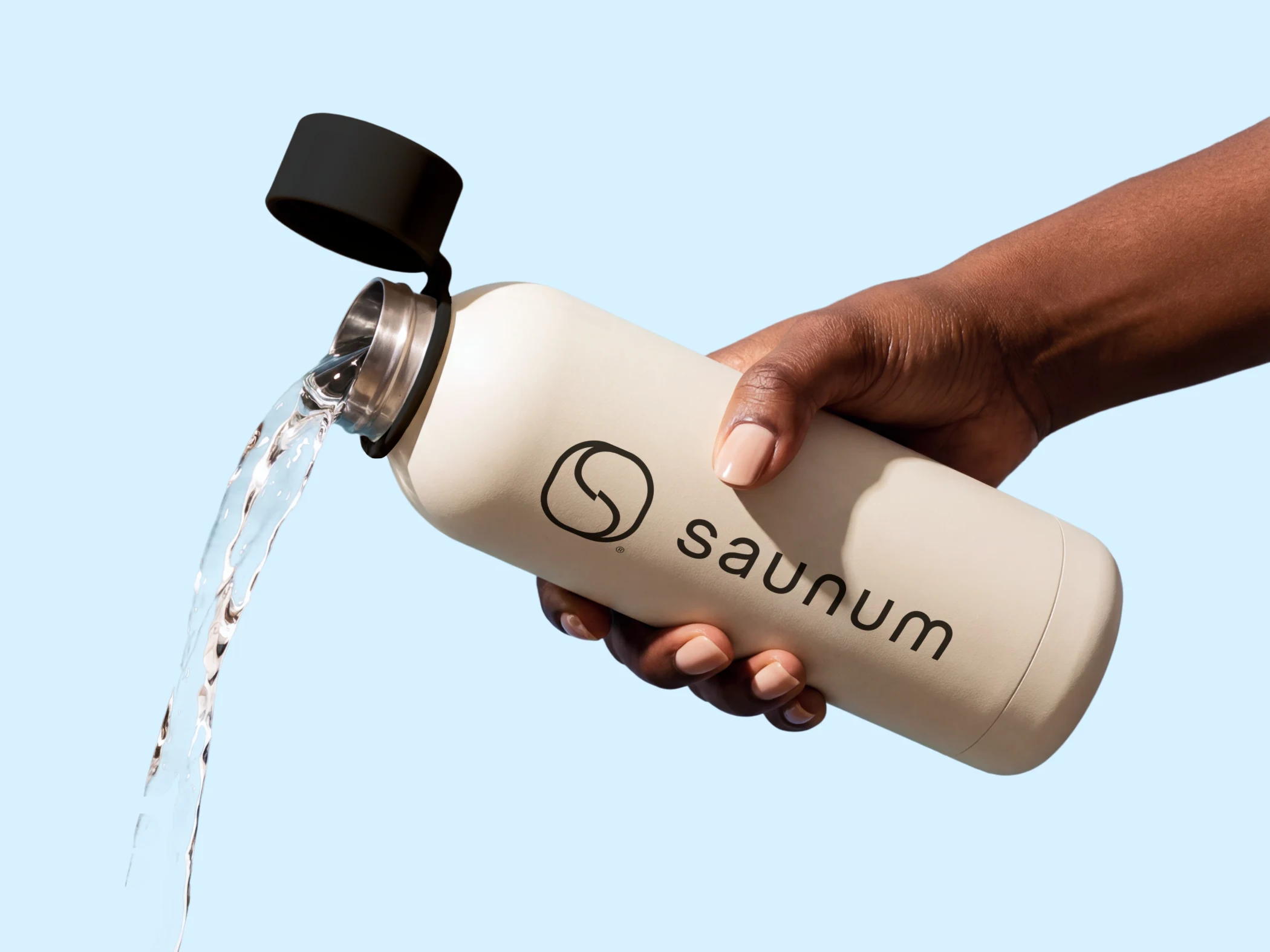 Hand pouring water from a beige Saunum water bottle with a black cap against a light blue background.