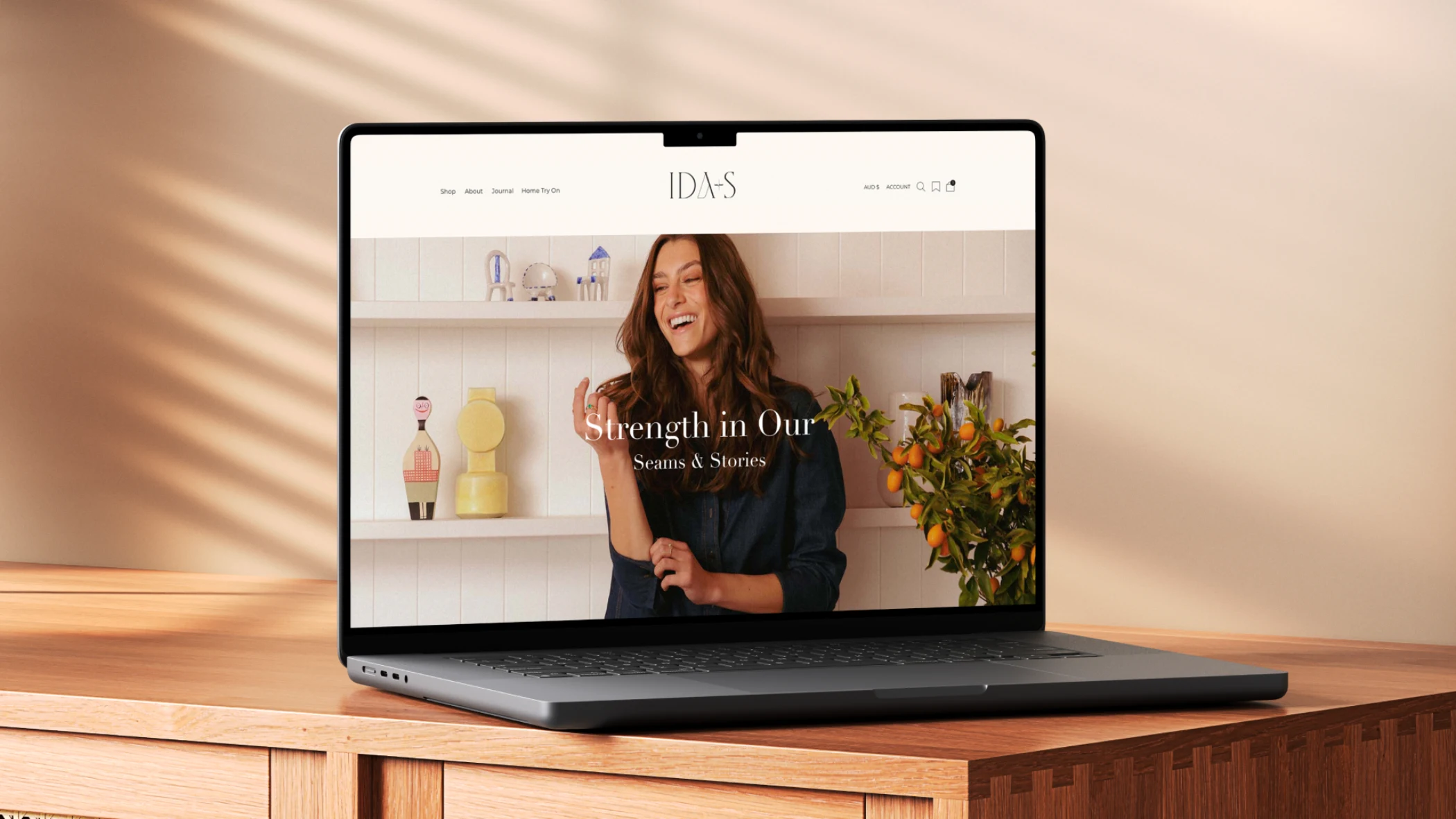 Open laptop on wood table displaying a website with a smiling woman and text 'Strength in Our Seams & Stories'.