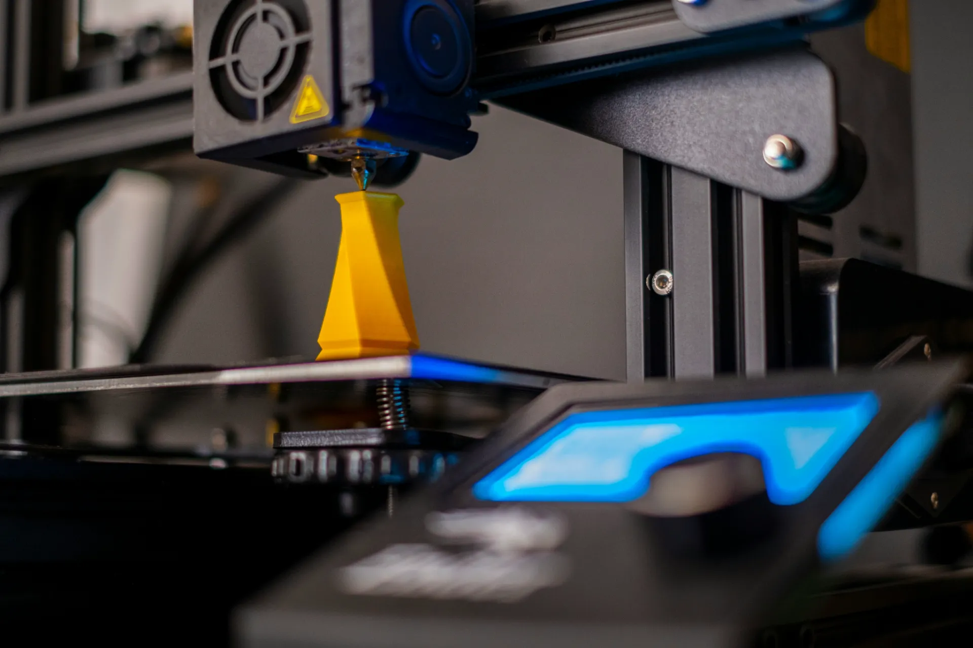 Close-up of a 3D printer printing a small yellow object on a heated bed with blue display controls.