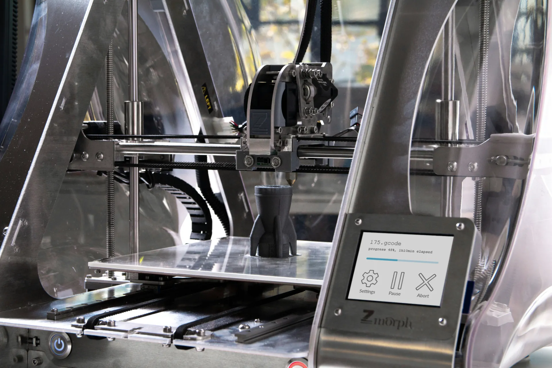 3D printer inside metallic frame printing a black object with progress displayed on screen.
