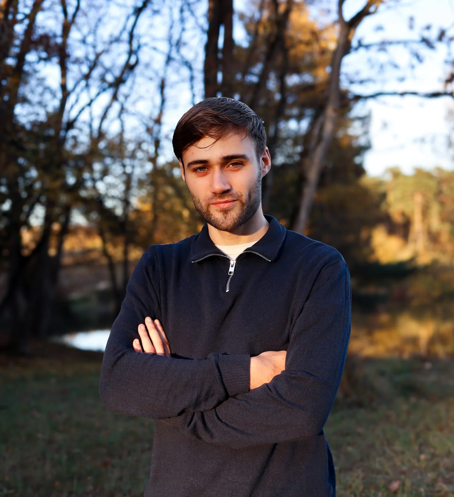 Young man with short brown hair and beard, wearing a navy sweater, standing outdoors with arms crossed.
