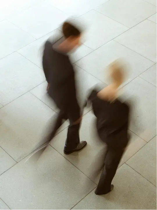 Blurred motion of two people walking on grey tiled floor, viewed from above.