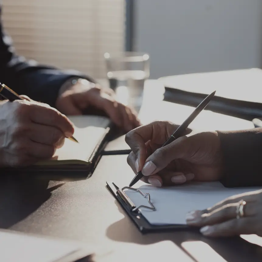 Two people at a table writing notes, one on a clipboard and the other in a notebook.