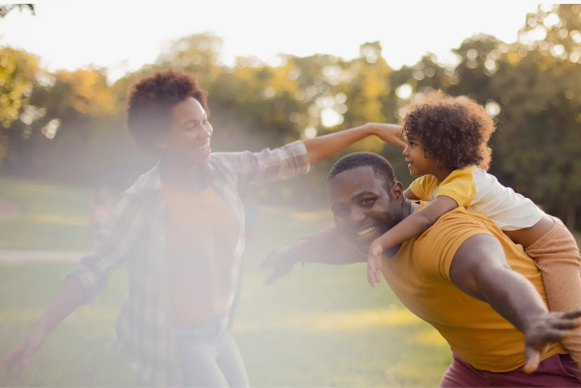 Smiling man giving a piggyback ride to a child while a woman dances nearby in a sunny park.