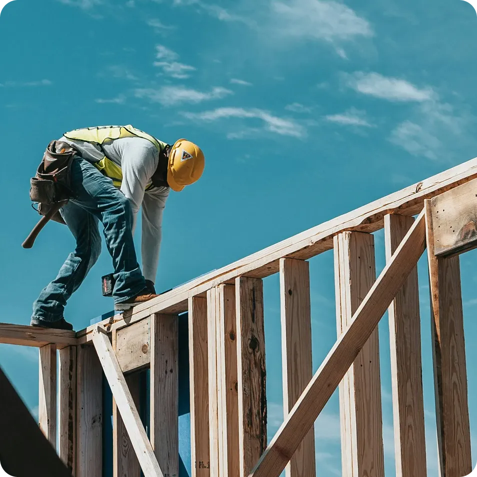 Construction worker wearing a hard hat and safety vest working on wooden framing under a blue sky.