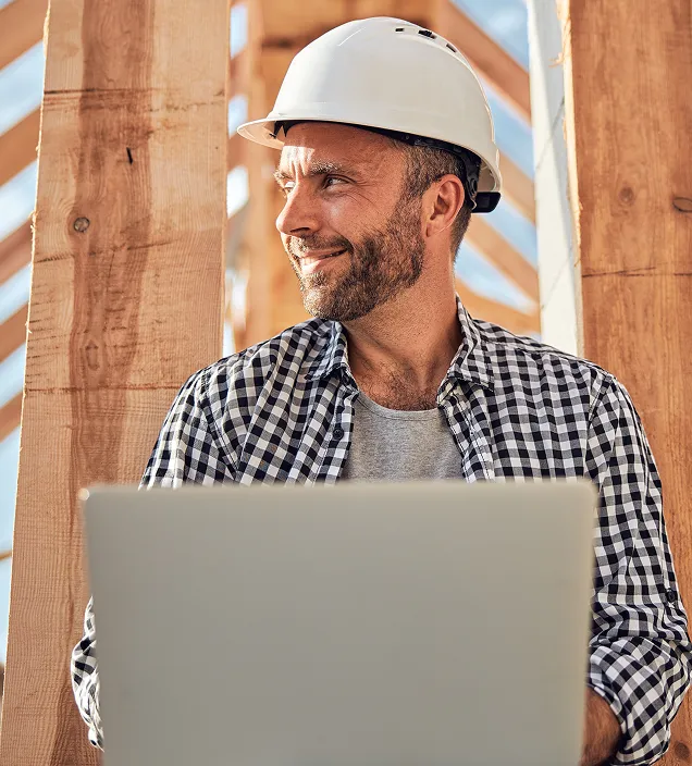 Smiling man in a white hard hat and checkered shirt holding a laptop at a wooden construction site.