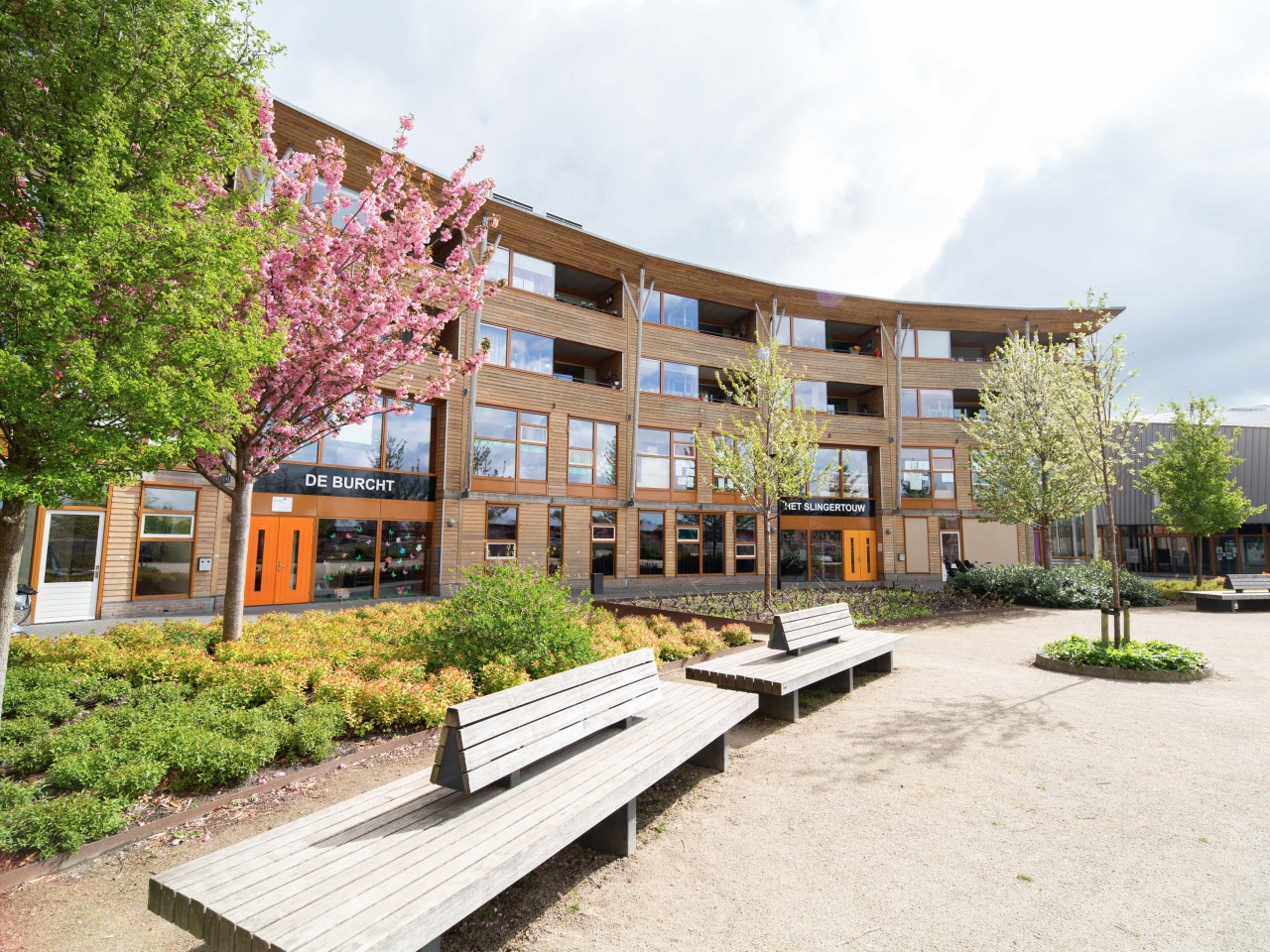 Gebogen houten gebouw met oranje deuren, bloeiende bomen en houten bankjes vooraan.